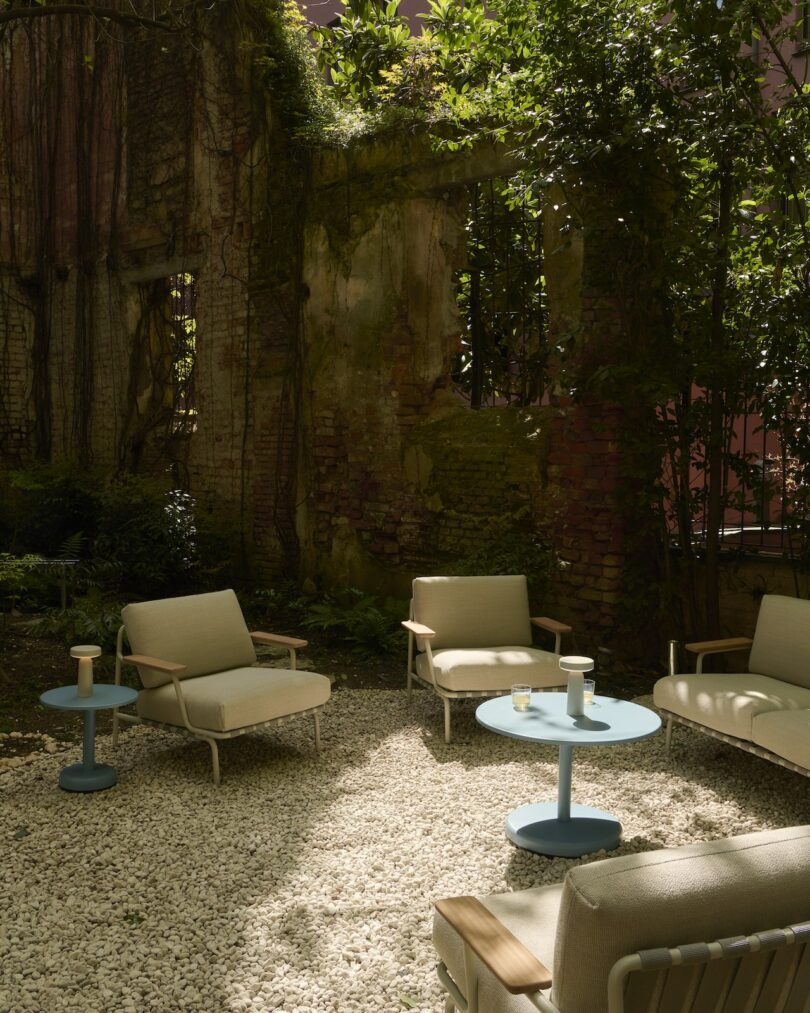 Four beige lounge chairs and two small blue tables are arranged on a gravel patio in a garden with overgrown greenery and partially ruined brick walls