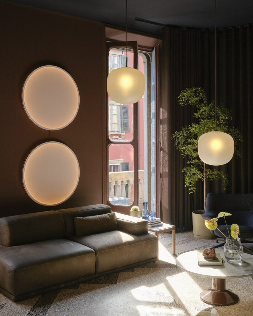 Modern living room with brown walls, a gray sofa, two round wall lights, hanging pendant lights, a round coffee table, and a green potted plant near a sunlit window