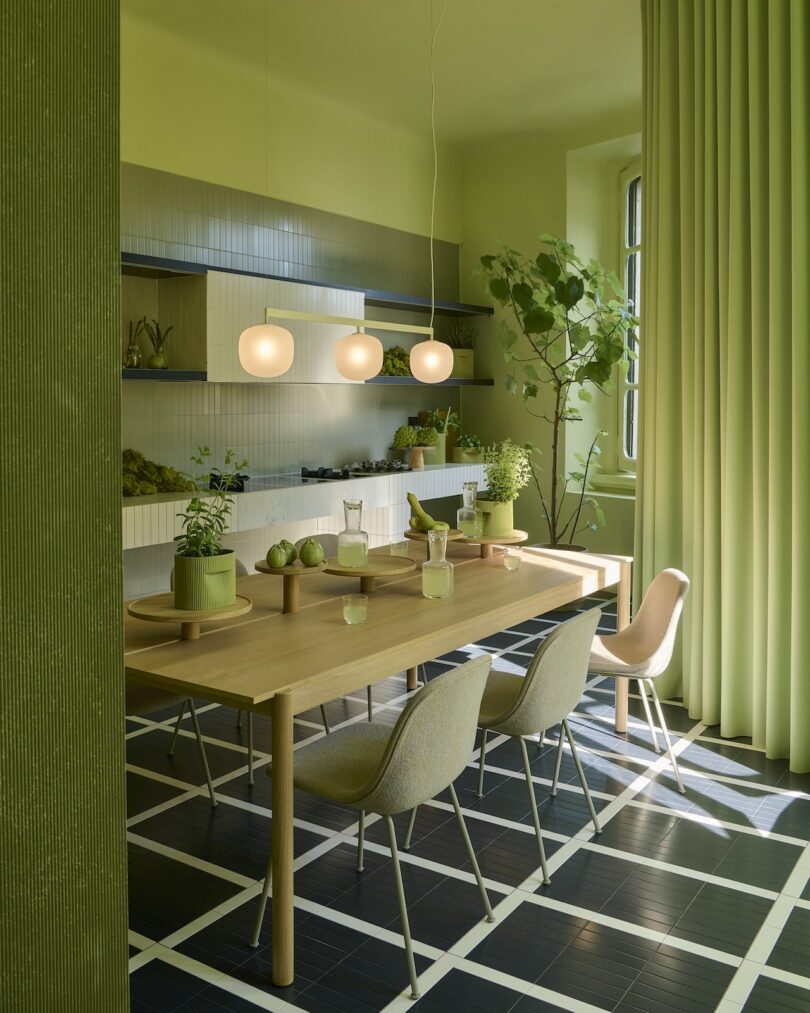 Modern dining room with green walls, plants, light wood table set for four, pendant lights, and large window with green curtains; checkerboard black and white floor tiles