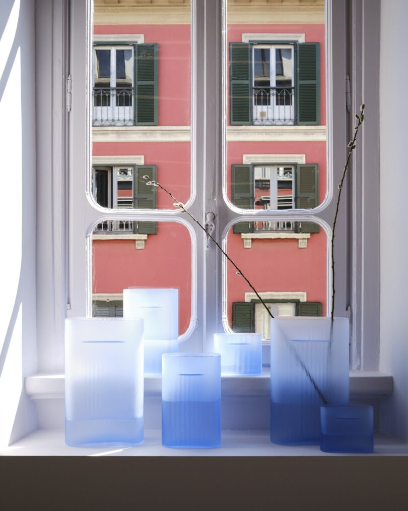 Frosted blue glass vases of varying heights are arranged on a windowsill, with a pink building and green shutters visible through the window behind them