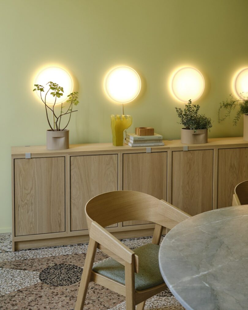 A light wood sideboard against a pale green wall with three round wall lights, potted plants, books, and a glass vase; a marble-topped table and wooden chairs in the foreground