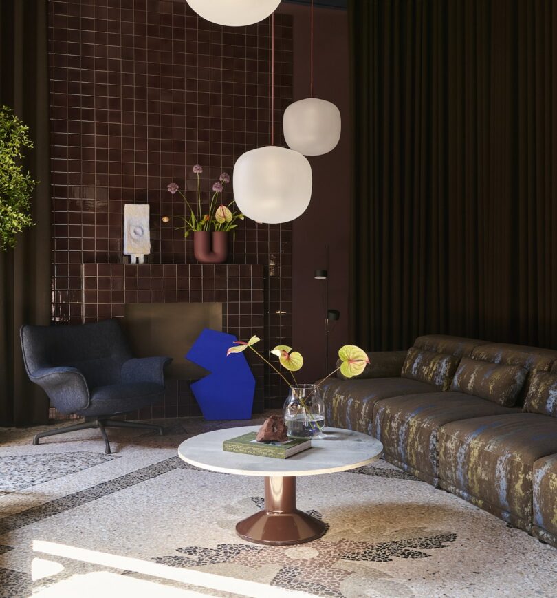 Modern living room with a brown tiled wall, sculptural lighting, dark sofa, armchair, round coffee table with books and flowers, and decorative plants