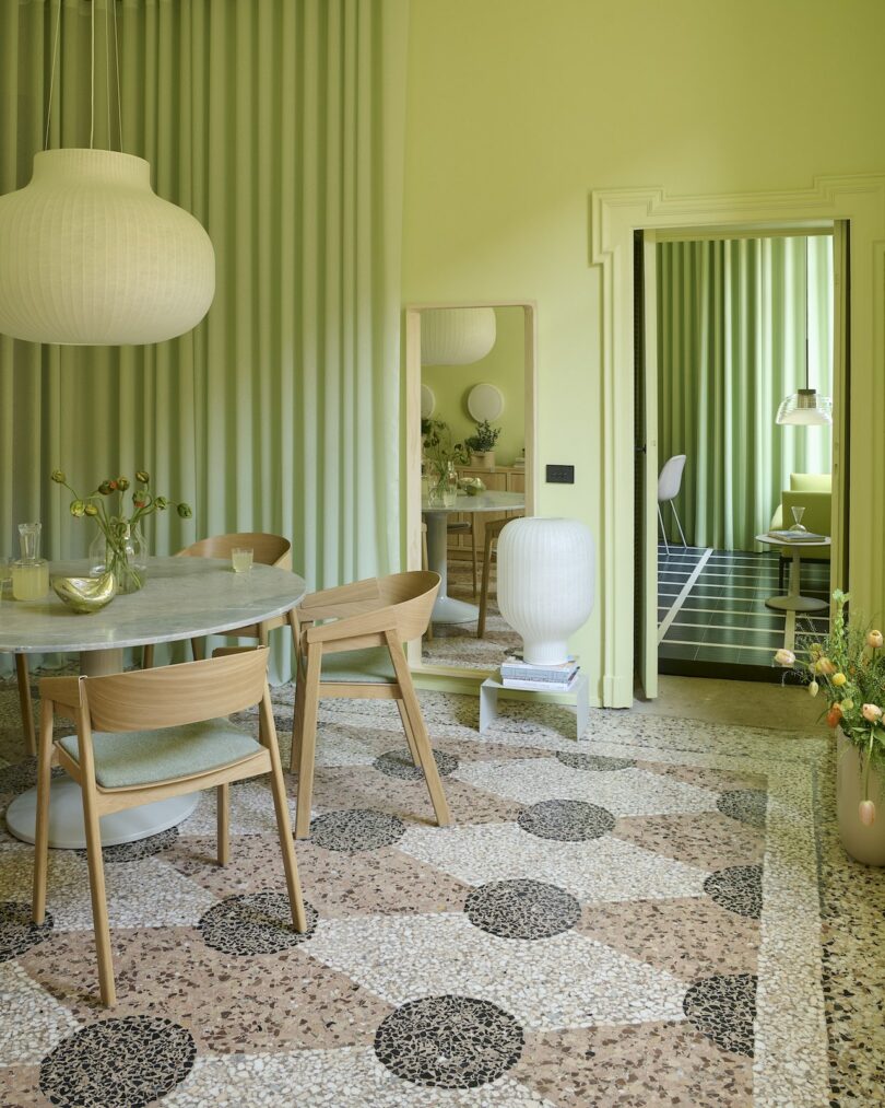 A light green dining room with a marble table, wooden chairs, terrazzo floor, round pendant lamp, and a mirror reflecting another green-themed room with a desk and chair