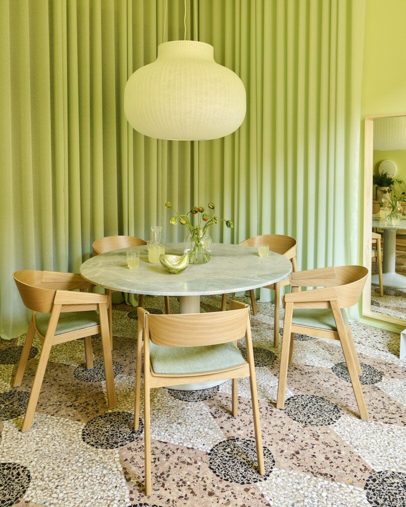 A round marble table with four wooden chairs sits under a large white pendant lamp; green walls, curtains, and terrazzo floor complete the modern dining area