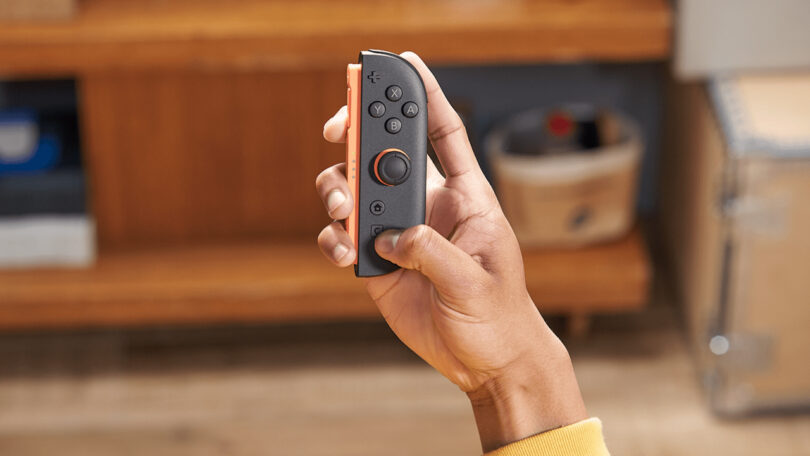 A person holding a black and red Nintendo Switch Joy-Con controller in their right hand, with a wooden shelf blurred in the background.