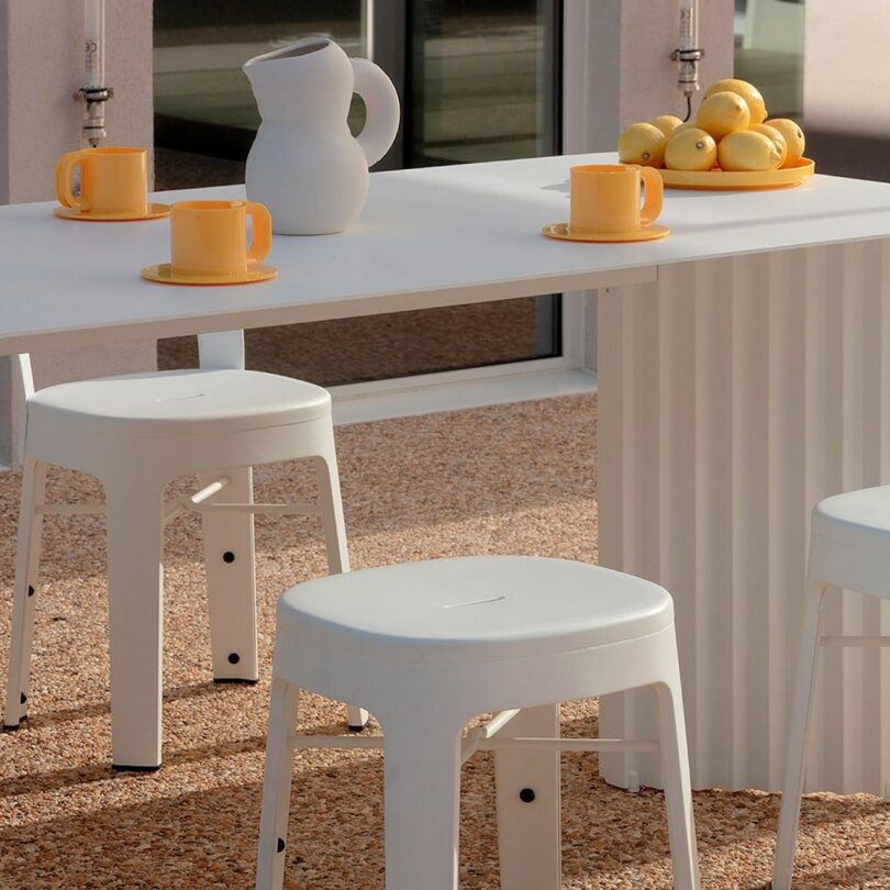 White patio table with yellow mugs, a white pitcher, and a bowl of lemons. Three white stools are arranged around the table