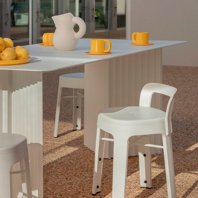 Outdoor patio with a white table and stools. A ceramic pitcher and four mugs are on the table. A bowl filled with lemons is at one end. Large windows reflect the surroundings