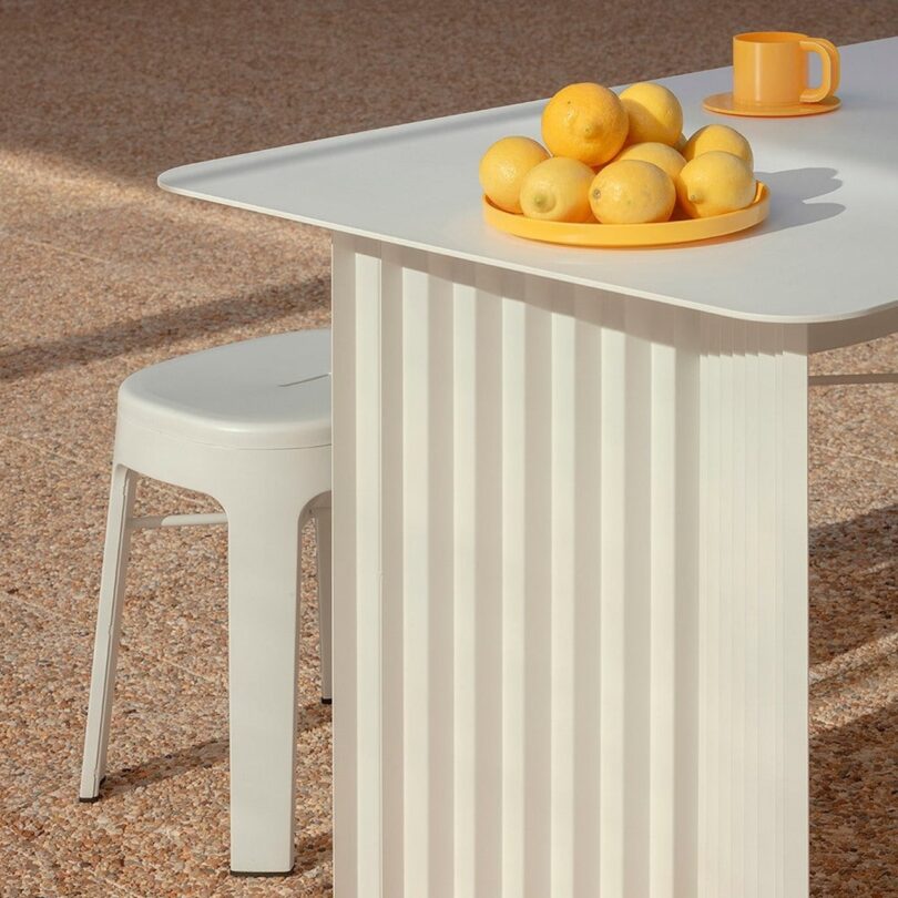 A white table with a fluted base holds a yellow bowl of lemons and a cup. A white stool is beside it. The floor is speckled beige and sunlight streams across the scene
