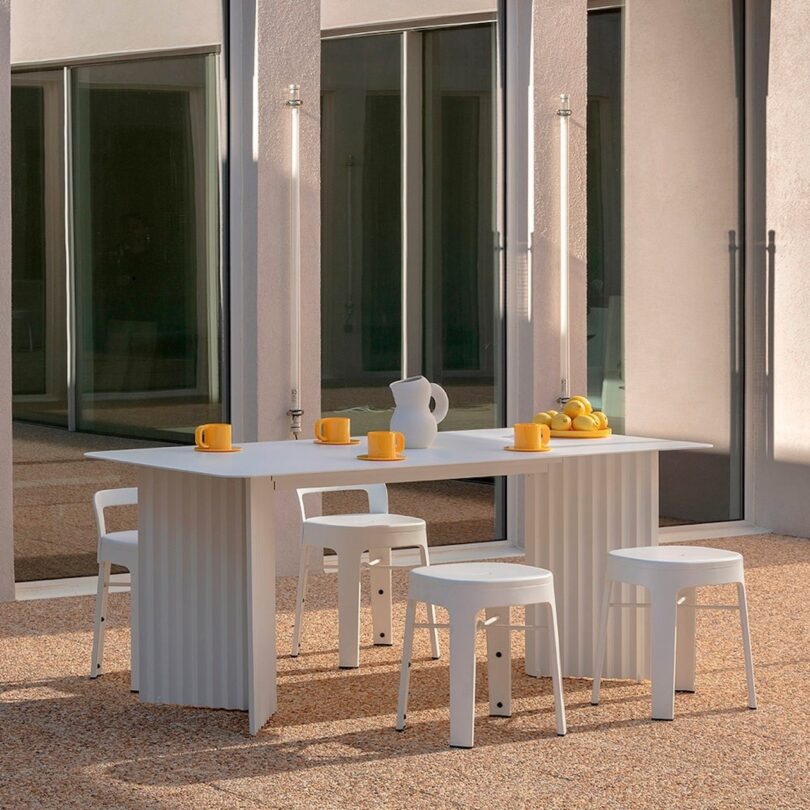 White outdoor table with matching stools, set with yellow mugs, a white pitcher, and a bowl of lemons. Glass doors in the background