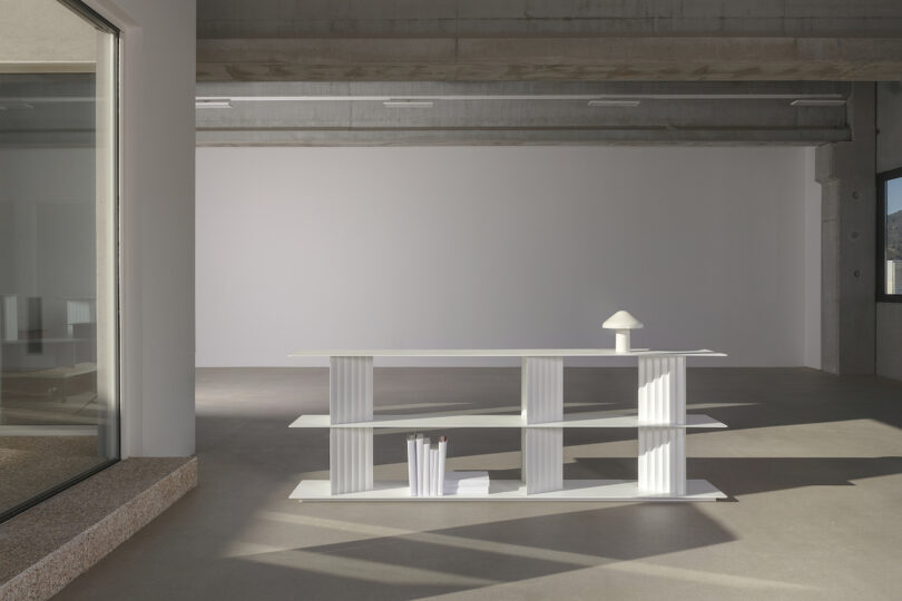 Modern white bookshelf with vertical supports stands in a minimalistic room. A small lamp and books are on the shelves. Large window on the left. Concrete ceiling and neutral walls