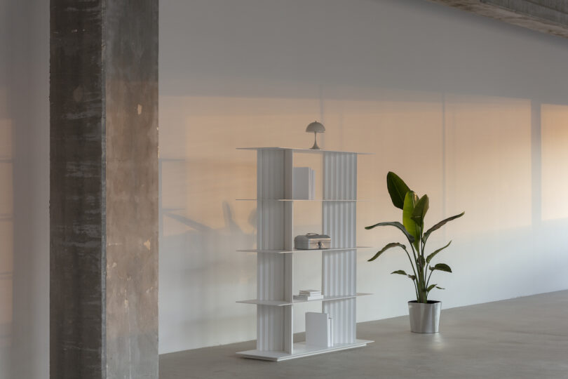 A minimalist white bookshelf with a lamp, books, and decor stands next to a potted plant in a spacious room with a concrete column and soft lighting