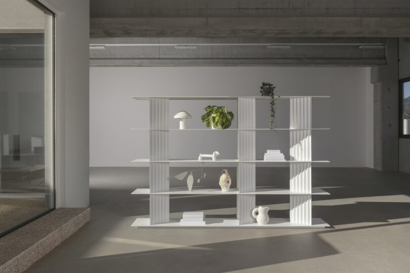 A modern, white shelf with various decorative items, including plants, ceramic vases, and books, stands in a spacious, minimalist room with concrete floors and large windows