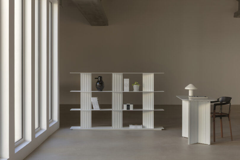 Minimalist room with a tall window, white bookcase displaying books and vases, a desk with a lamp and a chair, on a concrete floor