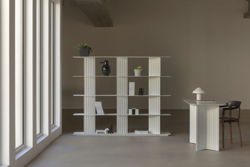 Modern minimalist interior with a white, open shelving unit and desk. Decor includes plants, books, and black-and-white objects. Large windows on the left let in natural light