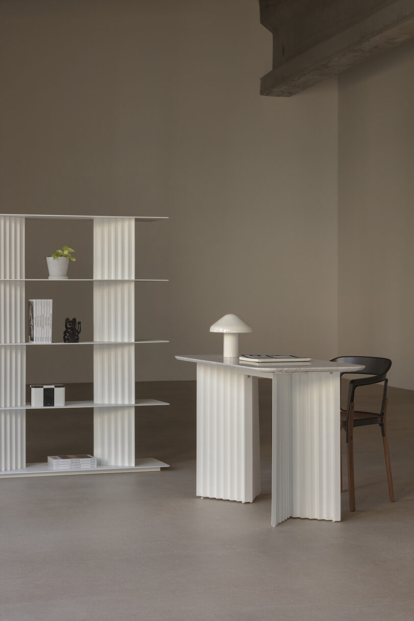 Minimalist room with a white fluted desk and chair. A lamp and books are on the desk. A matching bookshelf holds a plant, vase, and books. Concrete floor and beige wall complete the space