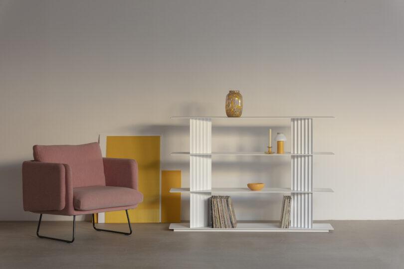 A pink armchair beside a white shelf with a vase, a bowl, and books. Two yellow rectangular panels lean against the wall