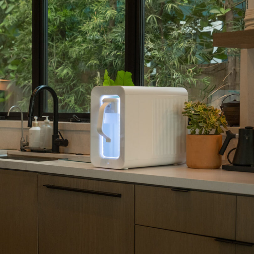 A white water purifier with a glowing blue light and sleek spout is placed on a kitchen counter next to a potted plant, with a sink and sunlit windows in the background.