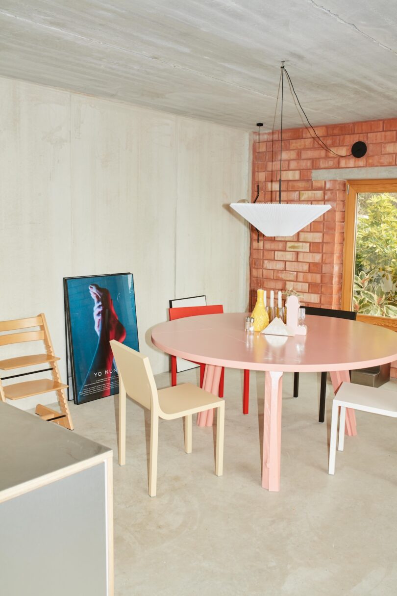 A round pink table with assorted chairs sits under a geometric pendant light; bottles and vases are on the table, with a framed poster leaning against a light brick wall.