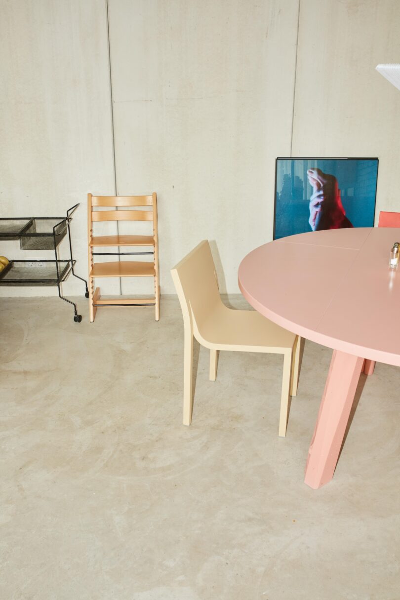 A minimal room with a pink round table, beige chair, wooden high chair, black cart, and a screen showing a hand on a blue background against a light-colored wall.