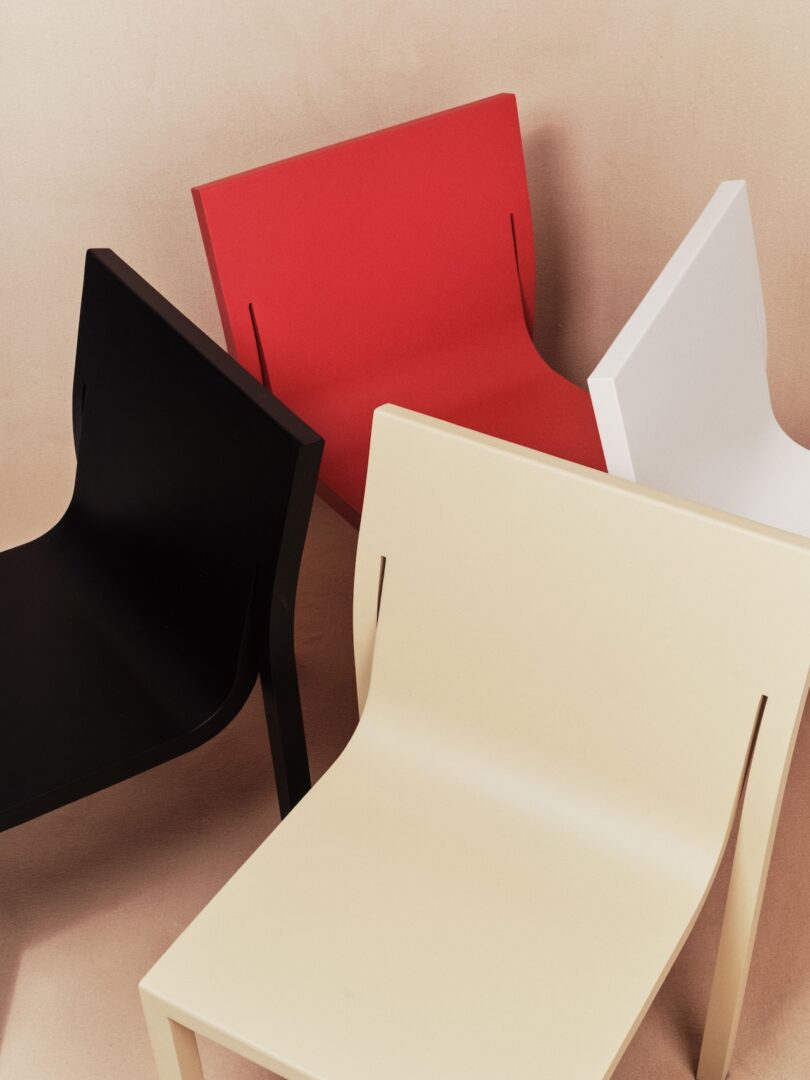 Four modern, minimalist chairs in black, red, white, and beige are arranged closely together on a light background.