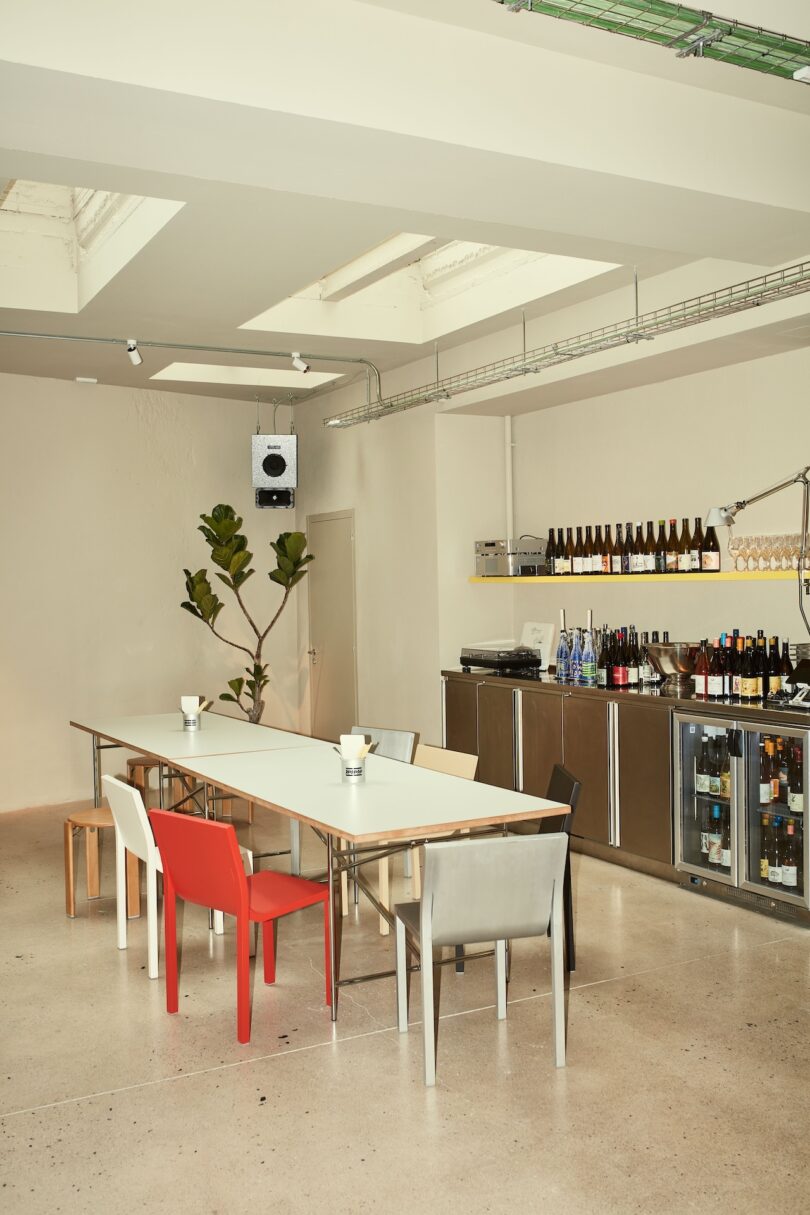 Minimalist dining area with a long table, assorted chairs, a potted plant, and shelves stocked with wine bottles above a stainless steel counter and glass-fronted refrigerators.