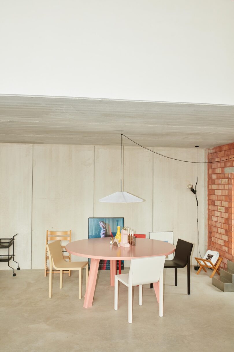 A modern dining area with a round pink table, assorted chairs, a pendant lamp, and decorative items against a minimalist wall with concrete and brick elements.