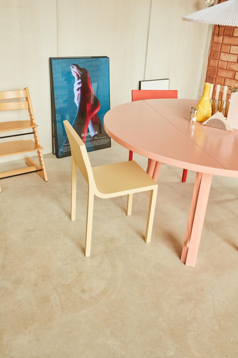 A yellow chair and round pink table are set on a concrete floor, surrounded by modern decor and a framed artwork leaning against the wall.