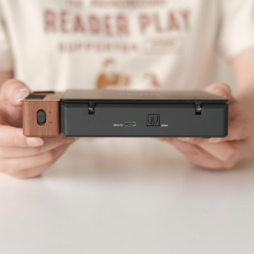 A person grasps a Syitren R300, a sleek black and wood-grain electronic device, its front panel dotted with various ports.