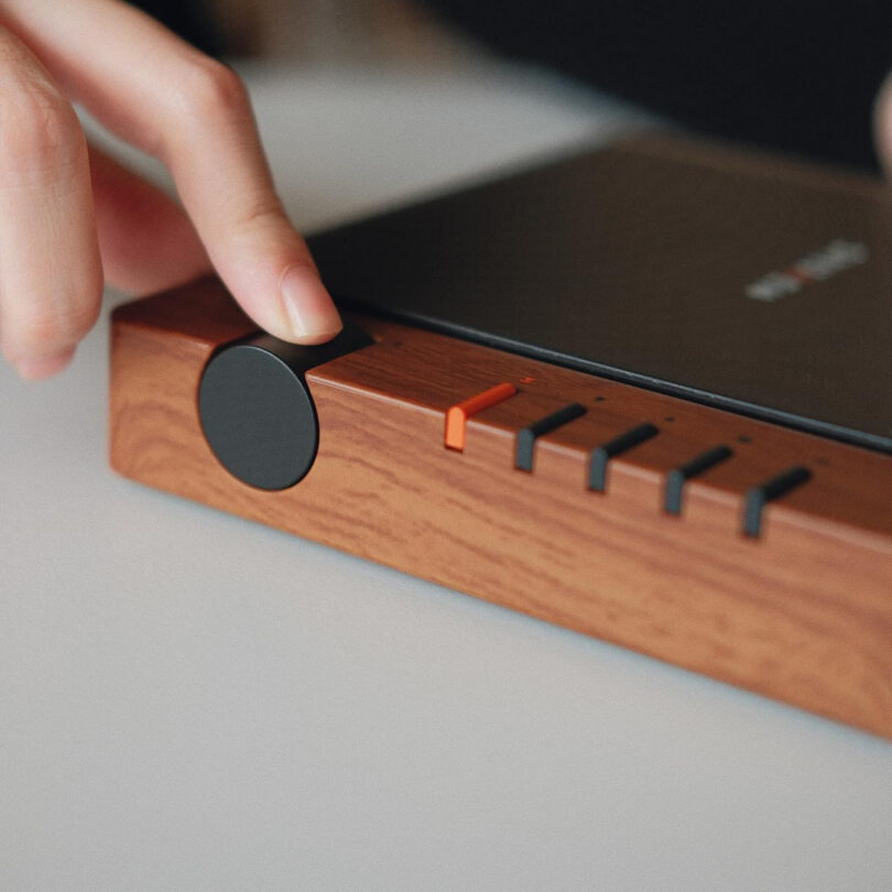 A hand adjusts the volume knob on the Syitren R300, a wooden audio device featuring sleek black and orange buttons.