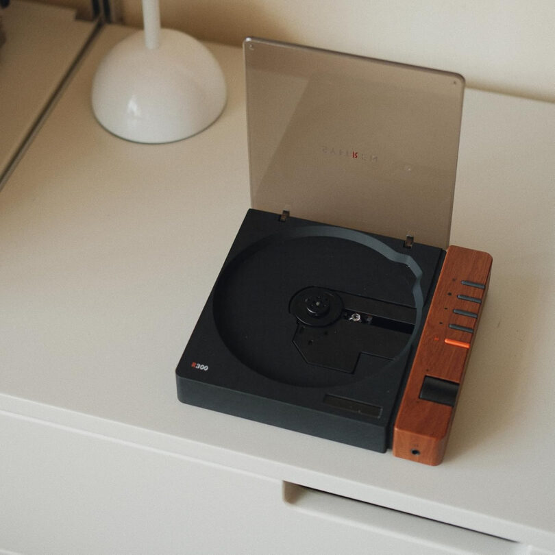 The Syitren R300 vintage turntable, with its wooden side panel and transparent cover, rests elegantly on a white surface. With the lid open, it reveals an empty black platform ready to bring music to life.