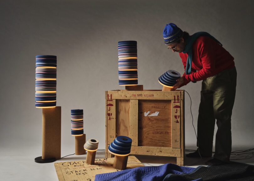 Person adjusting a Stackabl lamp with a colorful striped shade, next to a wooden crate labeled "Fragile." Other stacked lamps, some lit, are artistically arranged around