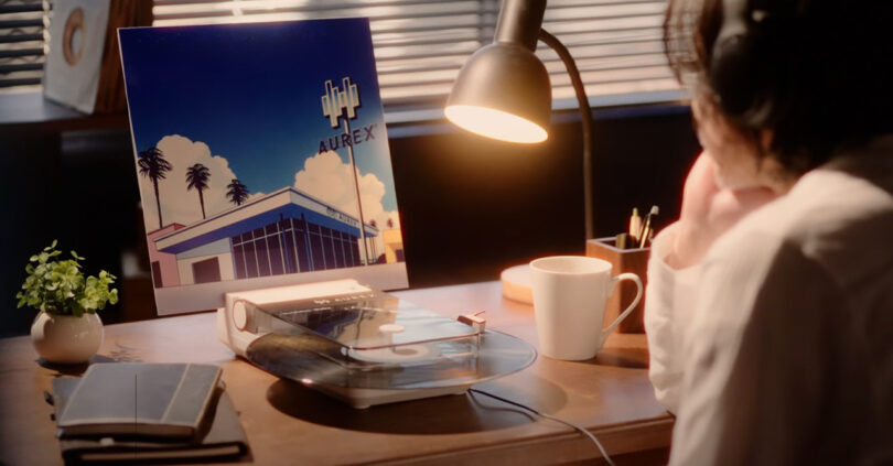 A person with headphones sits at a desk with a lit lamp, an AX-RP10 record player, a cup, notebooks, and a plant; an illustrated album cover stands in the background.