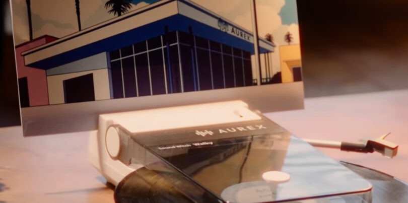 An Aurex AX-RP10 smartphone rests on a white device beside a power cord, with a photo of a blue building propped up behind it.