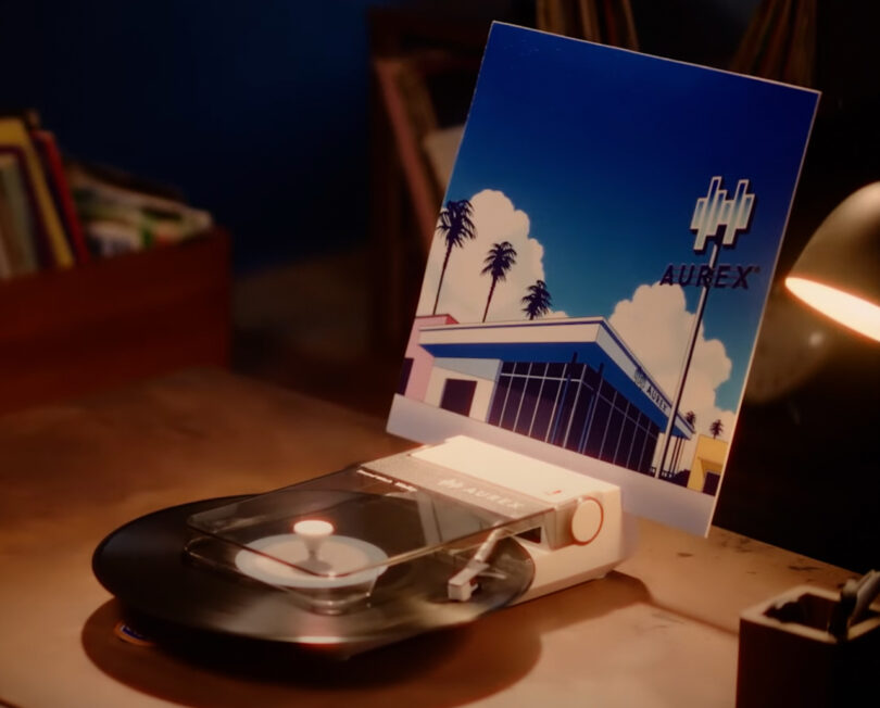 A vinyl record spins on a turntable, with its album cover—showing palm trees and a building labeled "AUREX AX-RP10"—propped up behind it on a wooden desk.