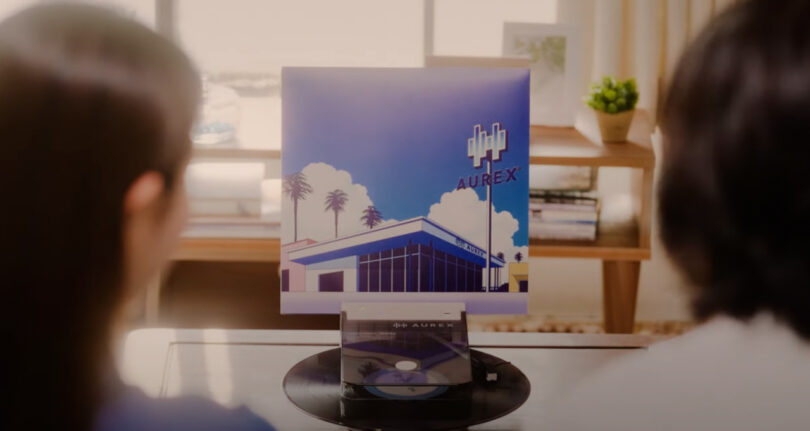 Two people sit facing a record player, the AX-RP10, with a vinyl album titled "Aurex" displaying artwork of a modern building with palm trees and a blue sky.
