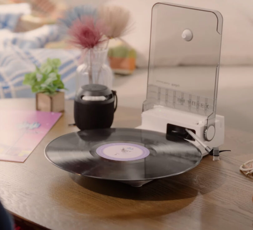 An AX-RP10 vinyl record player with a transparent cover is spinning a record on a wooden table, accompanied by a small potted plant and a black portable speaker.