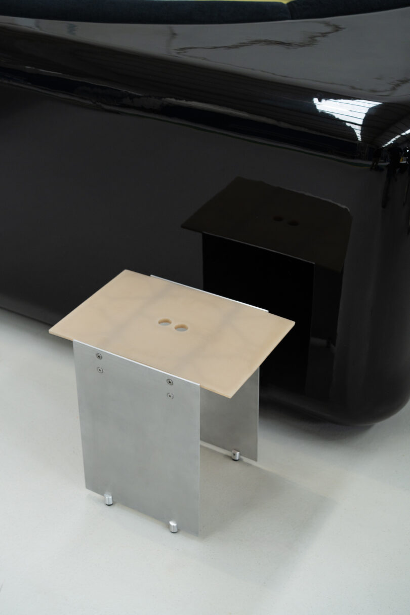 A small, modern metal table with a light-colored top stands on a smooth white floor in front of a glossy black surface