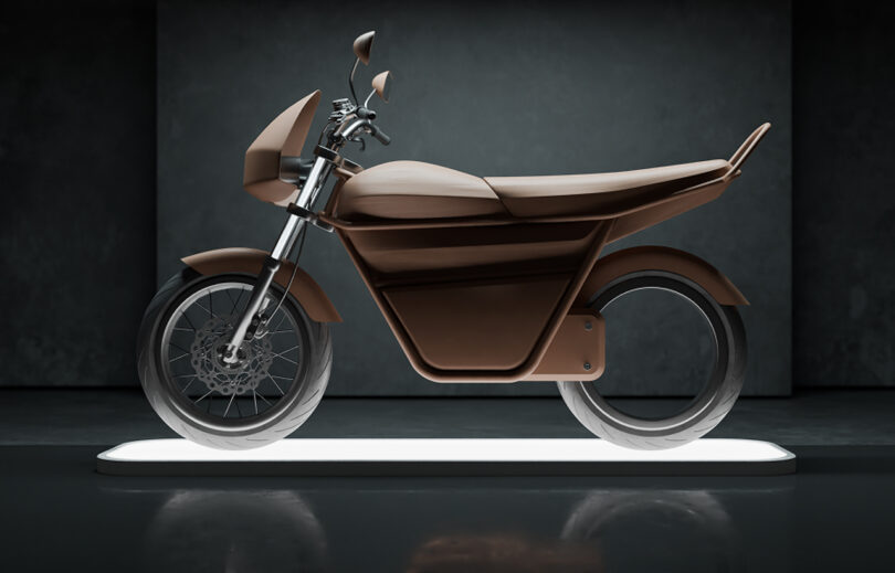 A brown, modern electric motorcycle is displayed on a white lit platform in a dimly lit room, photographed from the side.