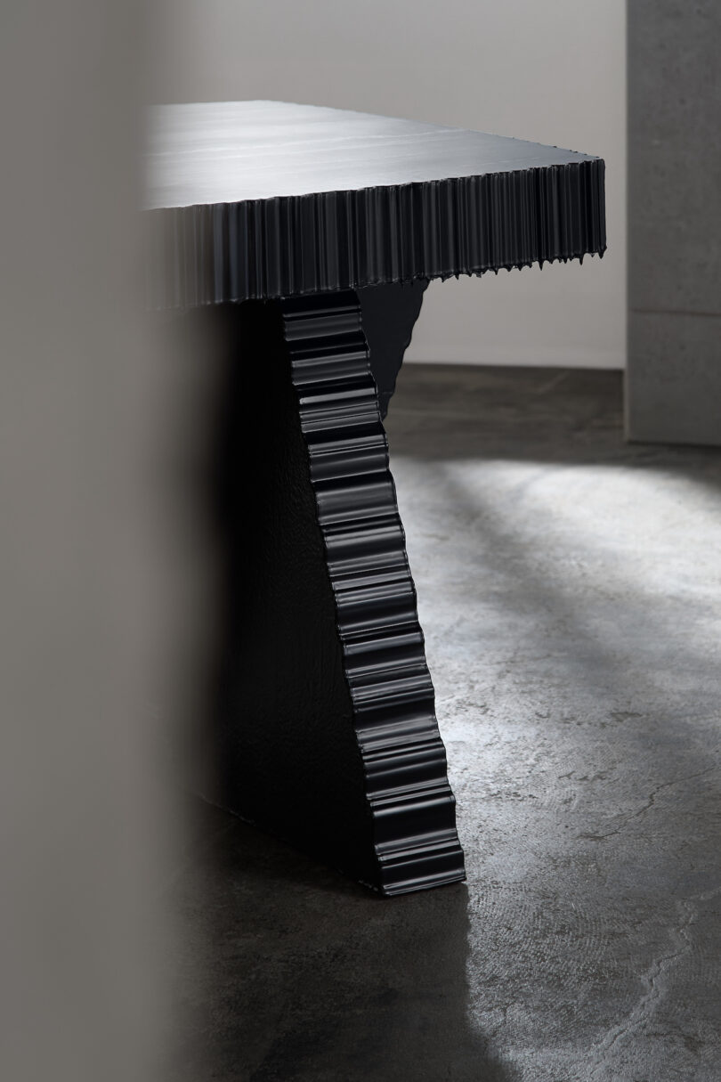 A modern table with a textured, ribbed design on the edges and legs, set in a dimly lit room with a concrete floor