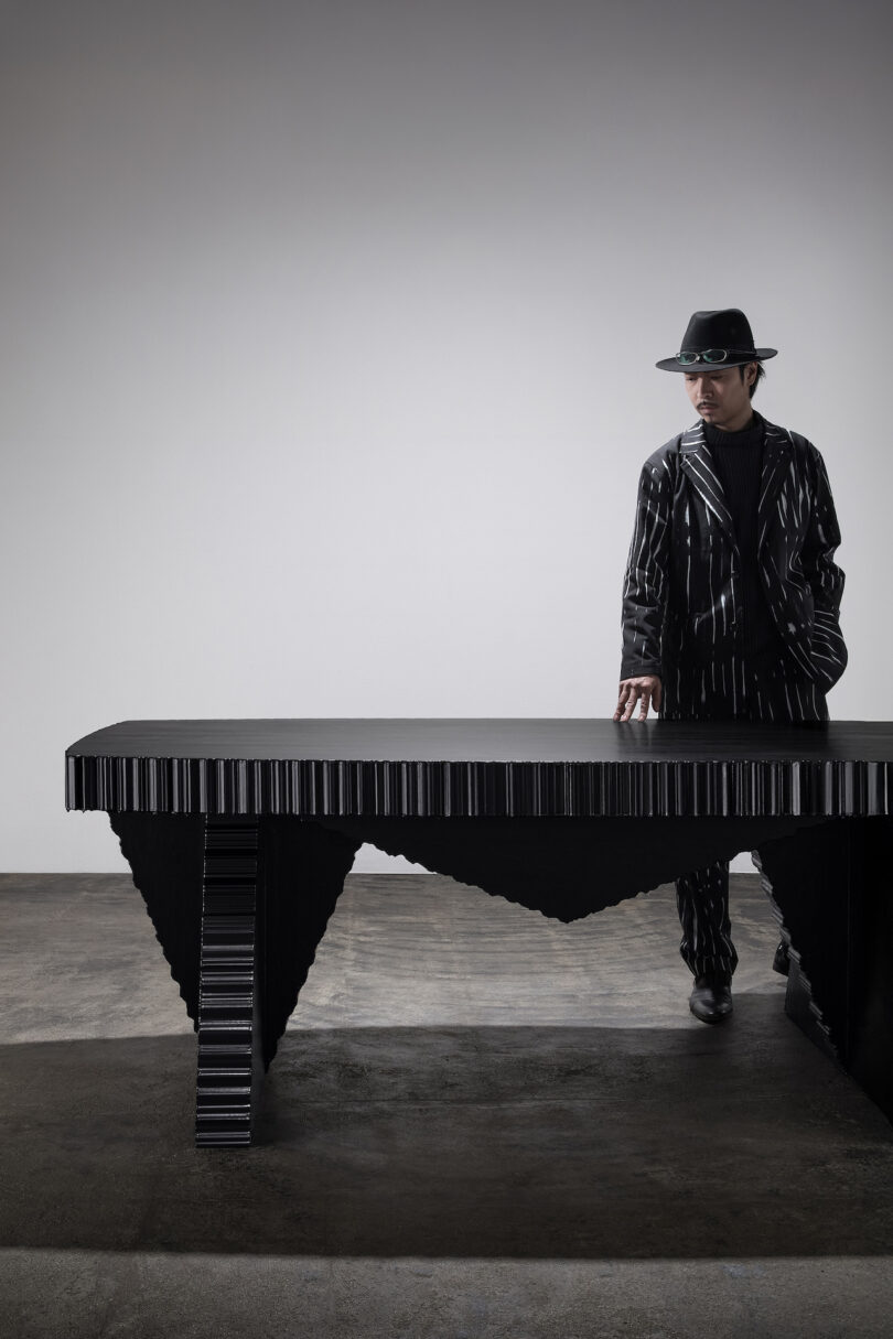 Person in a black pinstripe suit and hat stands beside a black, angular table in a minimalist setting