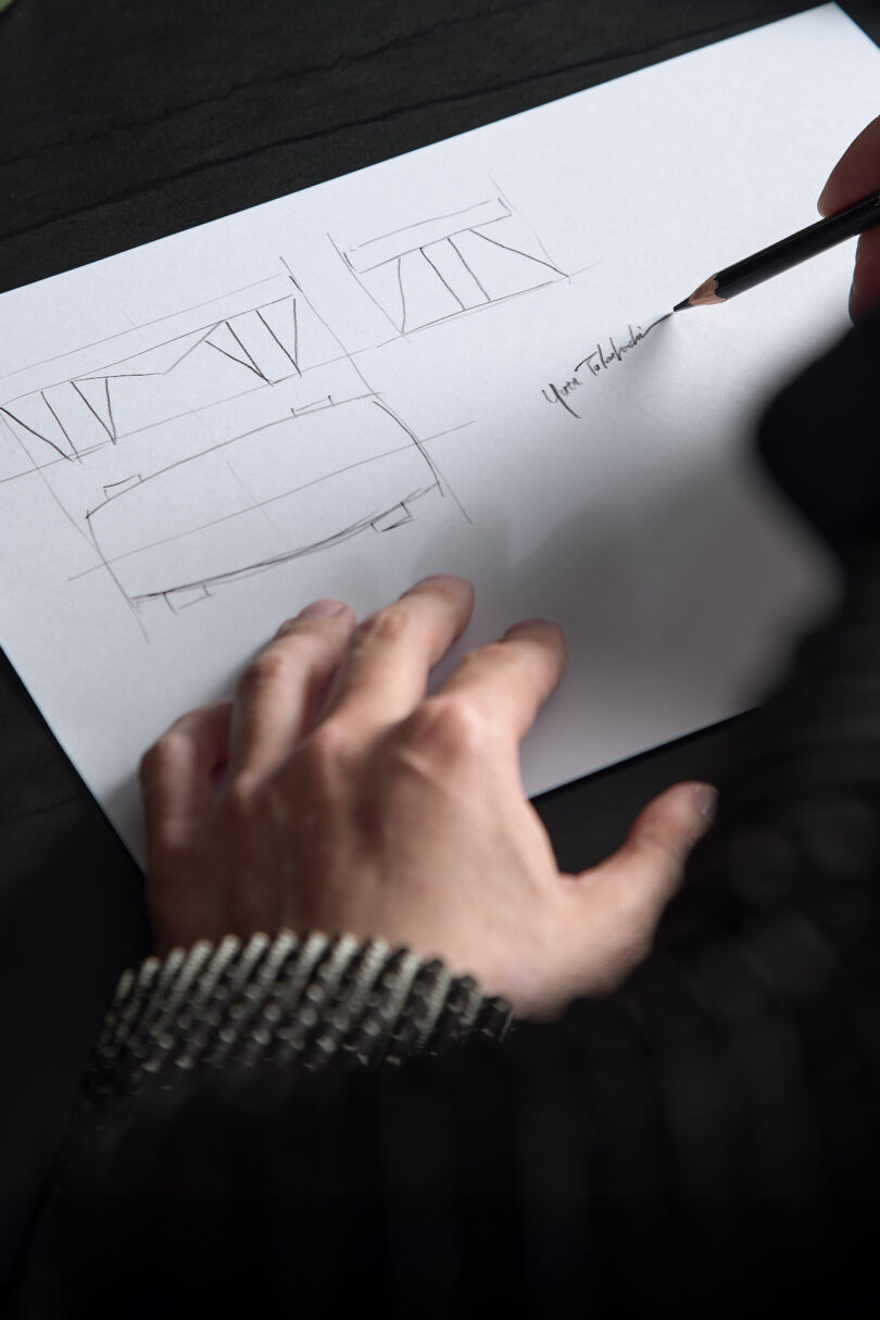 Person sketching designs on paper with a pencil, focusing on architectural elements and lines, on a dark surface