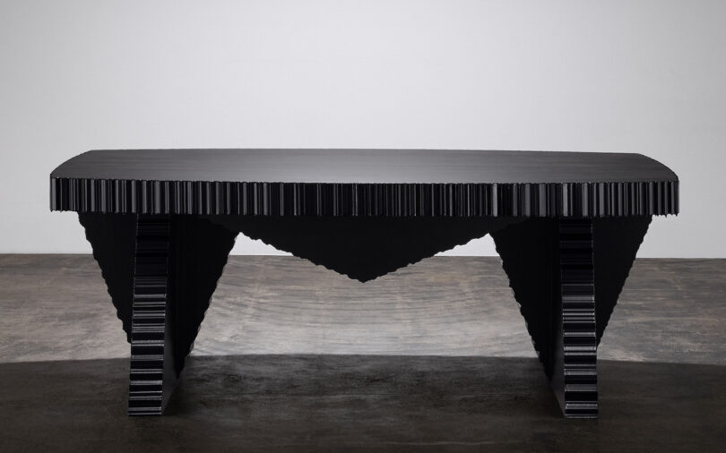 A modern, black sculptural table with a textured, jagged design on the edges and angular legs, set against a plain background
