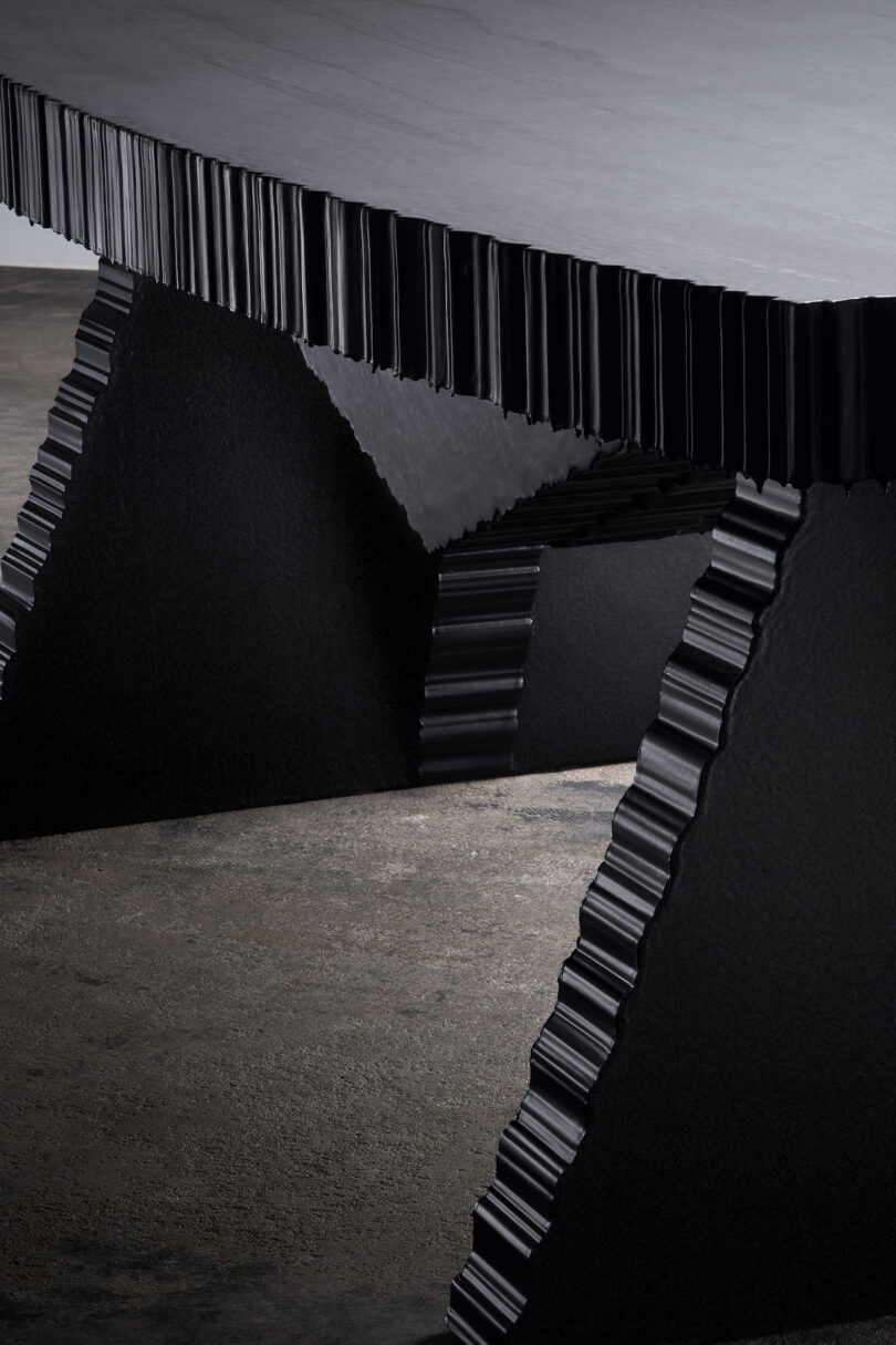 A close-up of a modern table with a textured, wavy black edge and angular supports on a concrete floor