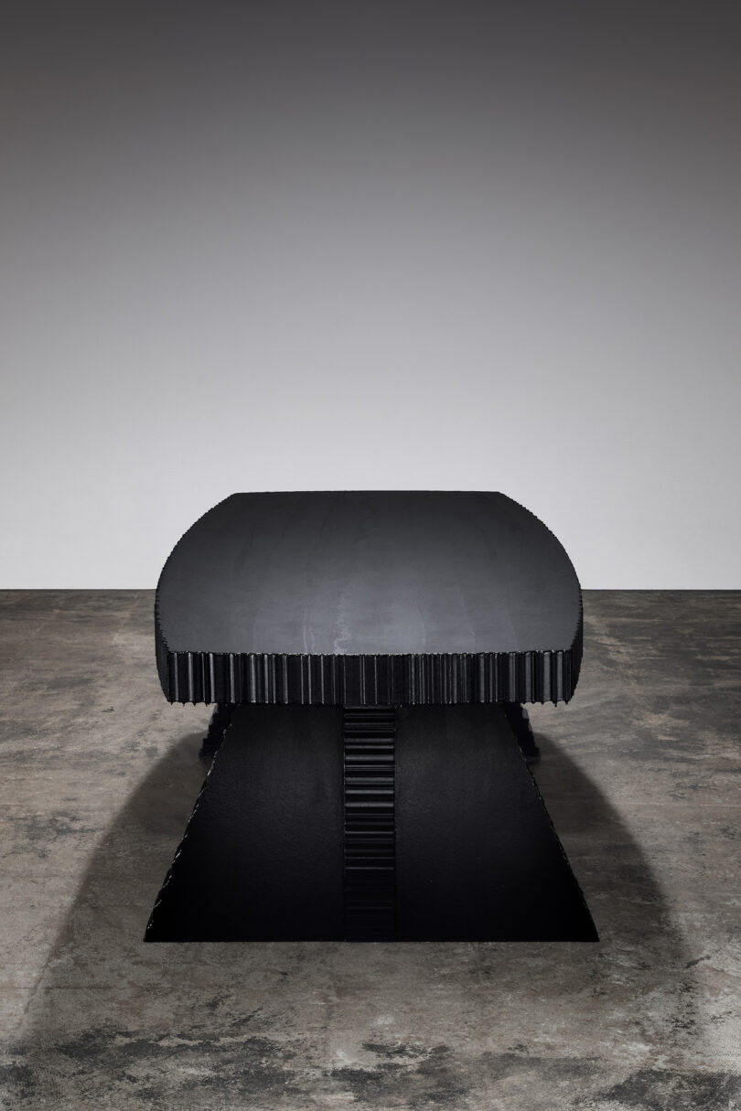 A modern black rectangular table with a ribbed surface, resting on a matching ribbed base, is placed on a textured concrete floor against a plain gray background