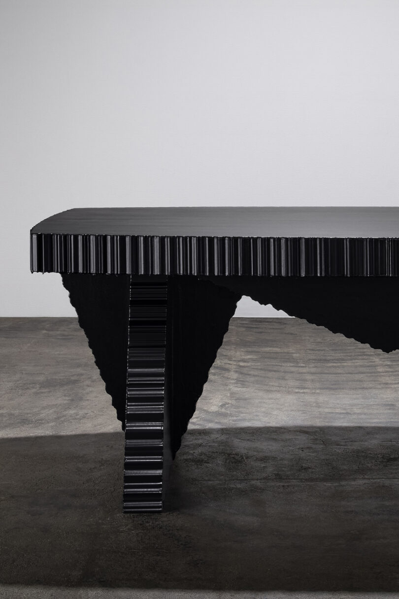 A black table with a textured, accordion-like edge and a triangular leg rests on a concrete floor against a plain white wall