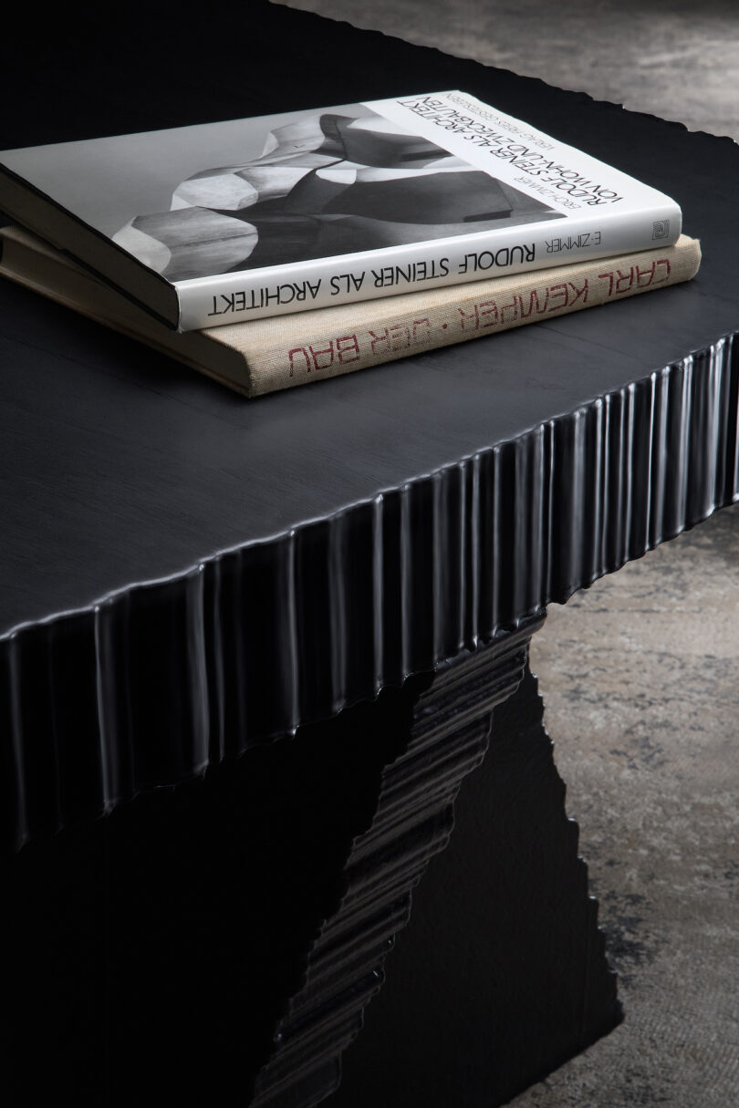 A black, textured table with two art books stacked on top: one open, revealing black and white photography, and another closed with red text. The surface reflects light subtly