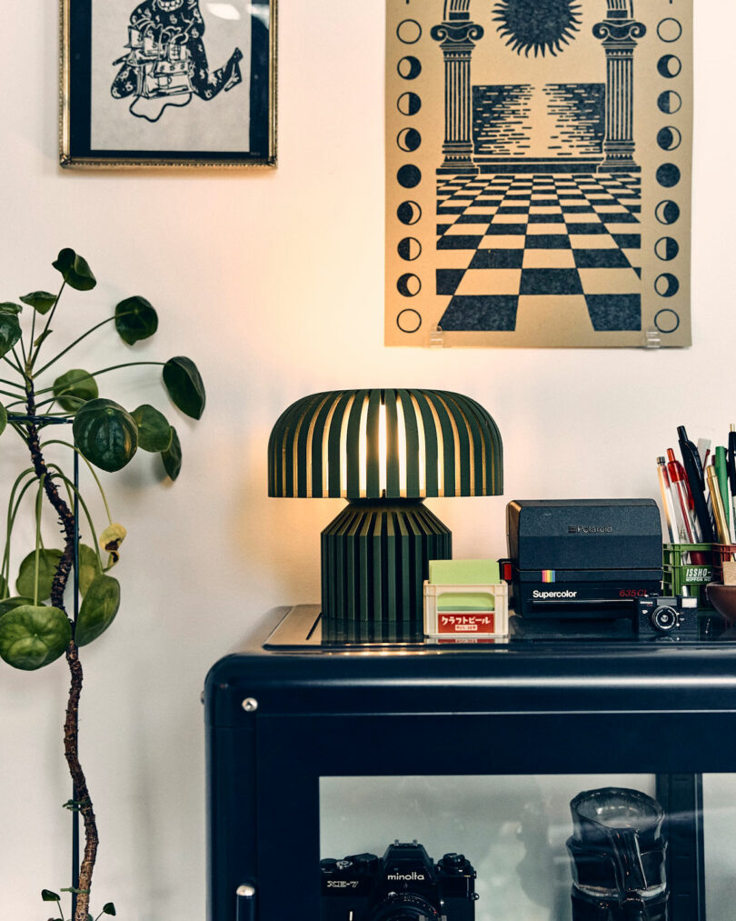A modern green table lamp by Walmen Dumaliang is lit on a black cabinet, surrounded by plants, cameras, pens, and wall art featuring graphic prints.