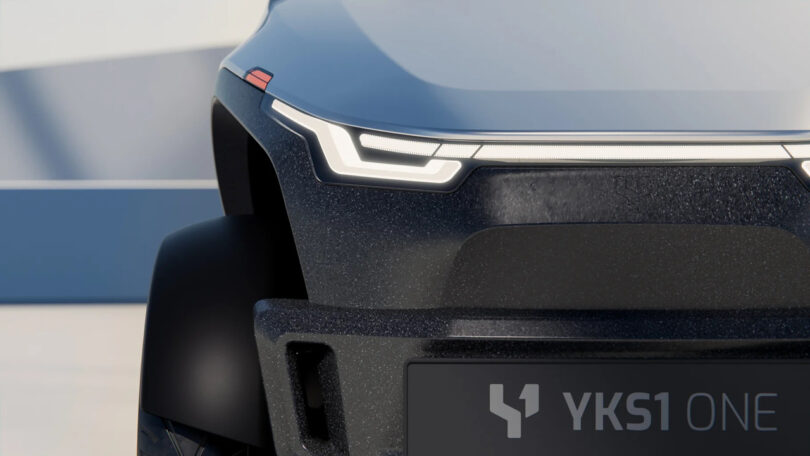 Close-up of the front of a modern vehicle, showing sleek LED headlights, a textured bumper, and a plate displaying "YKS1 ONE.