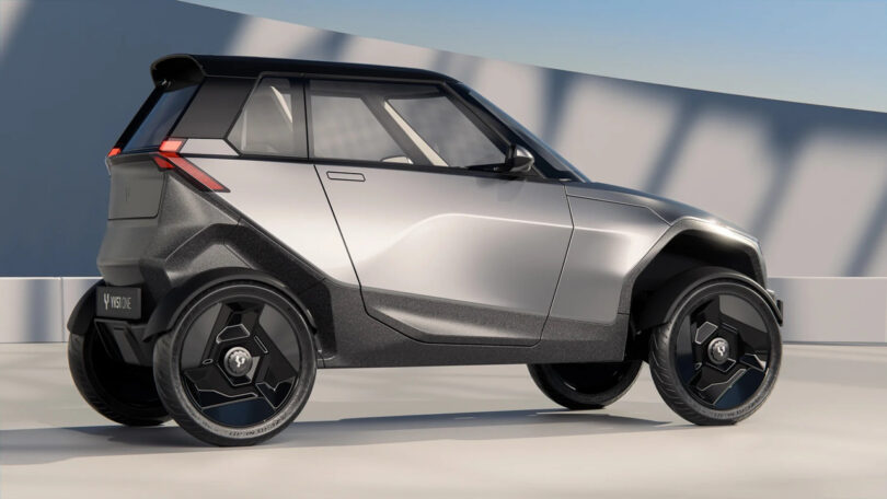 A compact, silver and black, futuristic-looking electric car is parked indoors on a smooth, light-colored floor with sunlight casting shadows in the background.