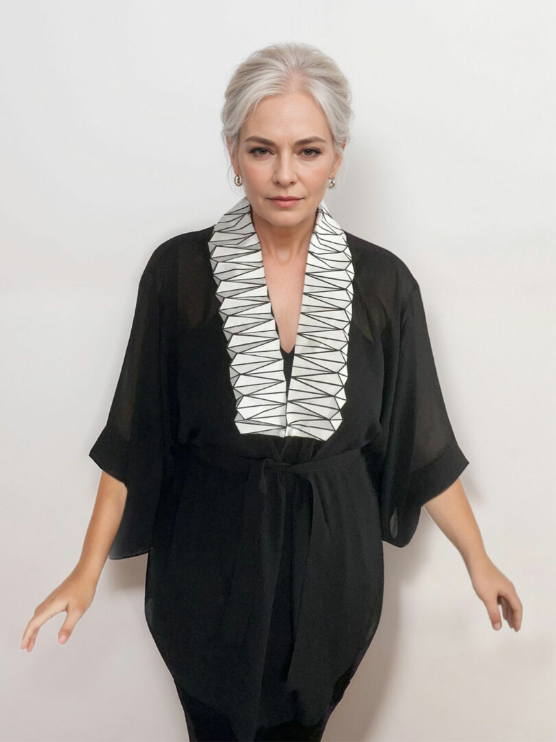 Una mujer con cabello gris recogido en un moño y vestida con un top estilo kimono negro con un cuello con patrón geométrico blanco de la colección 3Dtachables se alza sobre un fondo blanco puro.